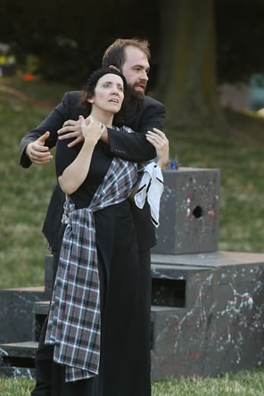Macbeth behind Lady Macbeth holds her to him as he talks. He's wearing a black jacket, she's in a black dress with a tartan sash. The multi-level platform is behind them, and a grassy hill and tree trunk beyond.