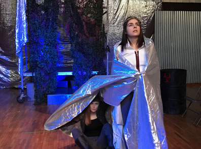 Lear in white jacket and black pants with a silver space blanket around her shoulders, and sitting on the floor the Fool underneath the space blanket next to Lear. In the background is a worker's rolling scaffold with camoflage curtain, a silver sheet backdrop, amo boxes, and a barrel