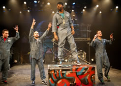 Pringle stands on a psychodelic painted trunk, the other three stand on the stage with their hands up, and the DJ is in the background on a scaffold