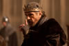 Henry IV, with fur cap and fingerless gloves, simple gold crown over a skull cap, points a finger up as he speaks with a threatening look