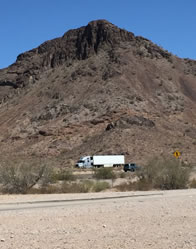 Photo of a rock-clif mountain surrounded by desert, with trucks passing on the highway at its foot
