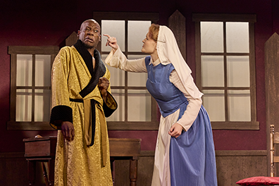 The Governor in a gold robe lined in brown fur winces as Susanna, in a blue and white novice nun habit, pokes her pinky at his face. In the background are windows and wood table and chair.