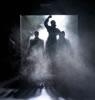 Coming through a large loading dock door with fog rolling in and a bight white spotlight from behind a woman in an overcoat, right fist raised, with other silhouette figures around her