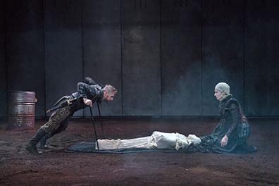 Production photo of Richard, leaning on his crutches over the white-shrouded corpse lying on the train of Anne's dress She is kneeling at the corpses' head, opposite Richard.