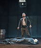 Production photo of Richard, in black unbuttoned Renaissance jacket and bround pants, leaning on his two crutches, standing by the shrouded body of Henry VI. The floor is dirt, a barrel is in the background, and a skull is suspended in a glass box hanging from the ceiling.