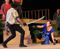 Mercutio in gold and white shirt, black pants and boots with a sabre in his right hand and sword in his left parries with Tybalt, in blue toga, black braw and yoga pants kneeling on the ground with a sword thrusting in her right hand and dagger in her left. In the background other characters watch on a sets of wood steps and balconies.