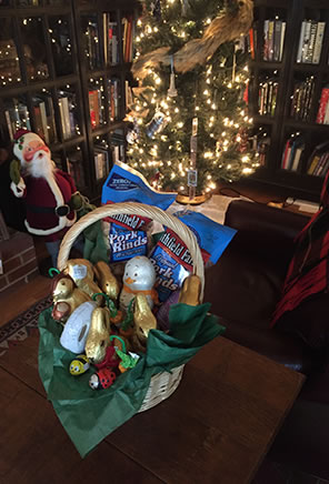 Photo of an Easter basket on a table with an Anna Lee doll Santa standing in the background next to a lit Christmas tree and book shelves.