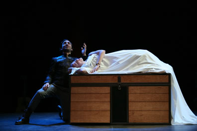 Iachimo in black kneels next to Imogen coverd in a white bed sheet on top of the trunk with a flat lid, a thick black vertical divider down the middle and plank lines on the two sections left and right of this divider