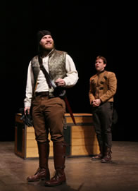 Cloten in brown pants and knee-high boots, silver vest, white shirt, sword hilt across his torso, Pisanio in the background in brown double-breasted waistcoat, brown pants, hands folded in front, the trunk behind him