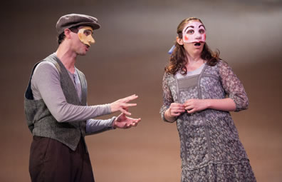George wearing brown dog cap, gray sweater vest, purple longsleeve under shirt and brown pants with hands motioning as he explains something, Emily in simple gray and purple print dress, a blue bow in her hair, looking out at the audience, her fingernails tentatively touching at her waist.