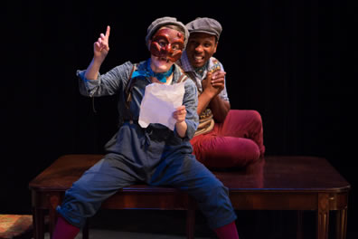 La Fleche reads a letter, held in his left hand as his right hand points up, and he's wearing blue overalls with suspenders and a blue work shirt and aqua scarf plus dog cap over a brown mask. Behind him, both sitting on a dining room table, Cleante in red pants and sweater vest over checked shirt and also in a dog cap, clasps his hands in joy. 