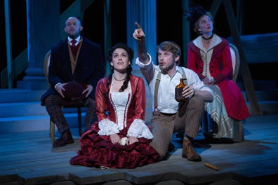 Jessica, wearing a red dress with ruffles below the waist and white breast and lacey sleeves, kneels on the floor looking up into the sky as Lorenzo next to her, wearing white shirt and tan pants with suspenders and holding a brown bottle of booze, points. Sitting on chairs behind them, Bassanio is in three piece suit with brown coat and checkered pants and wearing an overcoat and holding his hat while Portia wears a blue patterned formal dress with red riding cloack and a black hat 