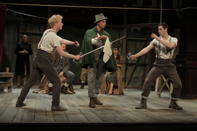 Hamlet and Laertes in farm pants and suspenders poise to duel with swords as Osric, in green cloak and hat, holds a handkerchief over the crossed swords