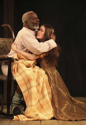 Lear in a white shirt is covered in a plaid blanket--you can see his stocking feet and bare leg beneath--sits in a chair hugging Cordelia kneeling and wearing a pattered brown royal robe.