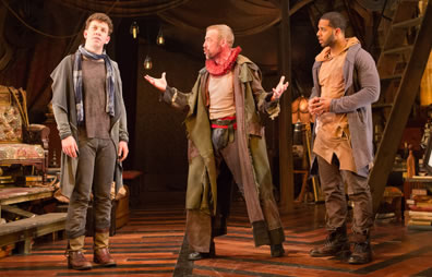 The Player with arms out and legs in a collosus stance talks with Guildenstern to his right, who is looking up and away, while Rosencrants on The Player's left looks at him. In the background is a stuffed chair behind Guildenstern and a attic ladder behind Rosencrantz and various junk. 
