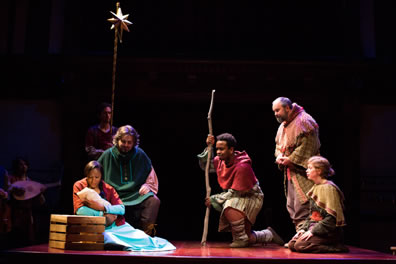 Mary ina  long blue dress sits next to a crate and cradles in infant in her arms, an orange shawl draped over her shoulder, Josephe in green cloak sits on a crate behind her, and across from them, Coll with his staff kneels on one knee, Gib stands with head bowed, and Daw kneels on both knees. An actor holds the gold Star of Bethlehem ona  pole in the background, and Kay plays a lute.