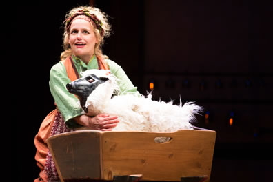 Gill in simple green shirt and orange dress with a red-patterned apron, hugs the sheep in a wooden cradle.