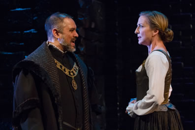 Burleigh, close-cropped hair and beard, black coat, a chain of office arond his breast, bares his teeth as he talks to Mary simply dressed in white, cuffed shirt, brown bodice, and brown skirt.