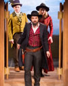 Gunslinging Petruchio in red vest and black overcoat and cowboy hat leas Kate in red riding coat and cowboy hat and Grumio in yellow buttler's tailes and feathered bowler through the saloon doors