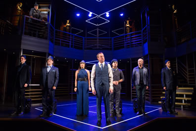 Timon in three-piece gray suit sans jacket stands in front of other cast members lined up on purple lightd stage and set, with Apemantus up on a balcony to the left.