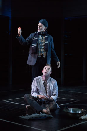 Timon sits on the floor in a dirty white shirt, pants, bare feet, a bowl next to him; standing behind him is Apemantus in casual jacket, checkered shirt, black and white striped scarf, and painter's hat holding up a tomato in his hand and speaking toward it