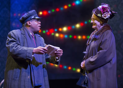 Hoke in overcoat and scarf and chauffeur's cap holds a small package in his hand as he talks with Daisy in simple overcoat with fur collar and hat featuring red and white flowers. Christmas lights in the background