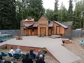 Photograph of the outdoor Fairbanks Shakespeare Theatre stage, with the wood facade Merry Wives set, ramps into the audience in chairs on the ground and platforms. Trees in the background