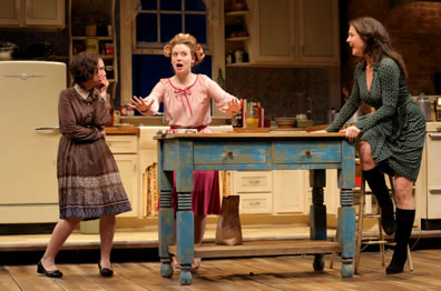 Lenny stands beside kitchen prep table in old fashioned brown and blue print dress, knock-kneed and hand to mouth; Babe behind the table, in curlers and pink blouse, her hands outspread as she talks; Meg sitting on stool at other end of table in low-cut green dress with black boots, hands on table smiling.