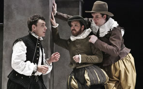 Hamlet in bulging black Elizabethan-style breeches, black vest and high collar white shirt to the left talks with Rosencrantz in green and gold breeches and jacket with white ruff collar dog cap and Guildenstern in gold pants brown jacket and lace collar with country brimmed hat, both leaning against a wall