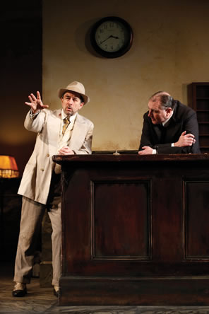 Erie in white sport jacket and fedora gestures with his hand as he talks, leaning on the hotel counter behind which is tne night clerk, hunged over, arms crossed, listening.