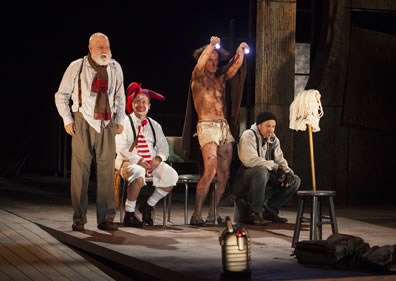 Lear stands in white untucked shirt, brown pants, suspenders, and red and brown scarf about his kneck, fool in white knee pants, socks, shirt, black suspenders, red-striped tie, red nose and white face with coxcomb sits, Edgar as Poor Tom with blanked around him and only in white cutoffs stands holding  up two flashlights, and Kent in black stocking  cap, gray hoody, fingerless gloves, and blue jean sits, all ecept the Fool looking at a mob stuck into a joint stool; Fool is looking up at Lear.