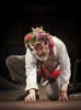 Lear, one knee on the ground, picking at the flor with his right fingers, wearing dirty white shirt and pants, his red regal sash, and the Fool's coxcomb covered in brambles
