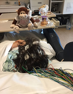 Photo of the back of Sarah in the hospital room, taken from the  head with multiple colored wires stringing across the pillow.