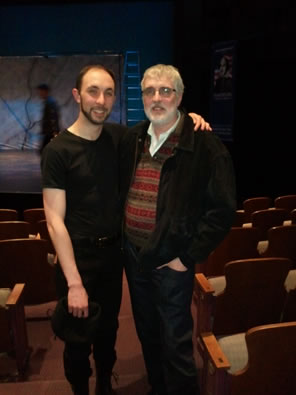 Jonathan in beard, slightly bald head, black t-shirt and pants with arms around Eric, gray hair and beard, glasses, brown and red striped sweater, black jacket and jeans, standing in the aisle of the theater