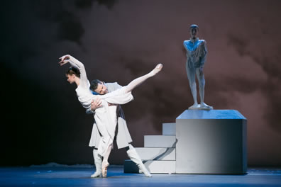 Hermione in white gown, on point with her right leg, left leg extended back and arms arching over her head, Leontes in white overcoat, arms wrapped around her waist and under her extended leg, head leaning into the crook of her back, a statue of Mamillius on a white pedestal in the background