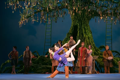 Perdita in a purple dress is in a flying pose, left leg extended back, right leg bent up, arms extended back, in the hands of Florizel in orange shirt and pants with blue smock. Other dancers and the Bansa stand in the background in front of the giant tree with long roots extending out to either side, two ladders disappearing up into the branches, and medallions hanging from the limbs.