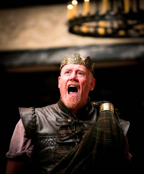 Macbeth in leather armor and crown on his head, mouth open in a shout with theater chandelier in the background