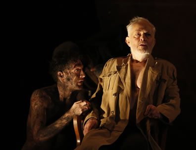 A begrimed Edgar looks up at Lear, shirtless but in trenchcoat sitting in a chair against a deep dark background