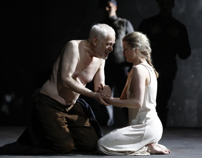 A shirtless Lear in brown pants, and Cordelia in white ankle-length slip kneel on the ground and clasp hands