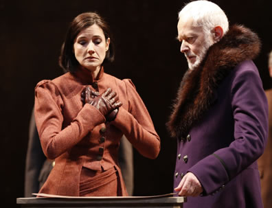 Regan in orange Edwardian dress suit and brown gloves holds her hands to her heart in a feigned expression of love as Lear, in fur trimmed purple cloak, shows her the map of her lands