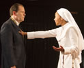 Isabella, in nun's white habit, speaks, smilingly, with left hand out (rosary beads hanging from the sleeve) and right hand on the breast of Angelo, wearing a dark suit and the gold chain around his neck