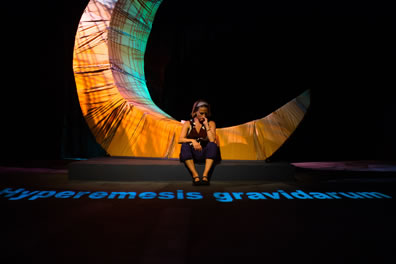 Jenn sits in front of the yellow crescent moon looking down at the words "hyperemesis gravidarum" projected onto the stage.