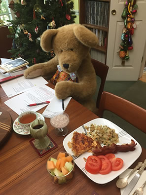 A big brown teddy bear sits at a dining room table, recipes spread in front of her, and a dish of food at the seat next to hers