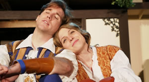 Orlando in blue and tan embroidered vest over white shirt, his leath-forearm-banned arm resting on his knee, Rosalind in floral gold vest on white shirt, the knees of her orange britches visible, and her head leaning on Orlando's shoulder.