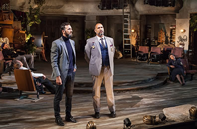 Production photo of Sebastian and ANtonio talking while the rest of the King's party sleeps in theater chairs.
