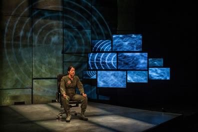 The Pilot in olive green flightsuit and boots sits in a chair with rollers, hands on knees, one with thumb up as if she's controlling a joystick, a bank of video screens behind her to the lest with images of a radar tracker over a dessert landscape, and a larger version of the same image projected on the wall behind her.