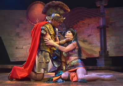 Antony in classic Roman legion uniform, skirt, leather armo, crest helmet and red ape on his knees stroking Cleopatra's neck as she holds  his arm, she sitting on the floor in a multi-colored sheen dress, halter, and bare midriff