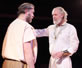 Timon in white nobleman's gown, gold medallion and wristbands talks gently with Flavius in simple shirt and pants and a headband