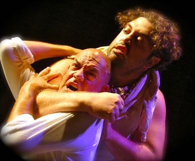 Curly haired, bearded Aufidius with bare arms has bald Coriolanus wearing a white T-shirt in a headlock