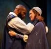 Othello with bald head and beard and blue cape embraces with Dessdemona in cap, blue cape and jewelled gown